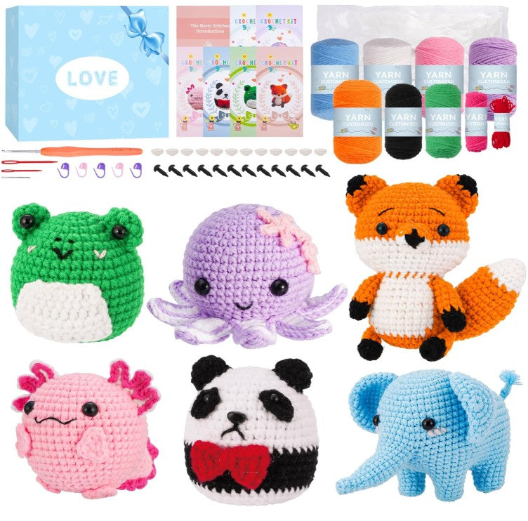 A crocheted amigurumi kit display showing finished handmade plush toys and yarn supplies: a green frog, purple octopus, orange fox, pink axolotl, black-and-white panda, and light blue elephant, alongside yarn skeins, small crochet hooks, beads, and a blue gift box labeled LOVE.