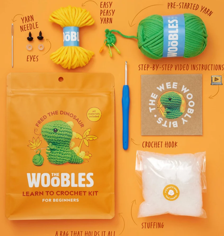 The Woobles Learn to Crochet Kit for Beginners laid out on an orange background, including a yellow yarn ball, green yarn ball, crochet hook, stuffing bag, yarn needle, pre-started yarn, instruction booklet, and branded pouch. Hand-drawn callouts label each item.