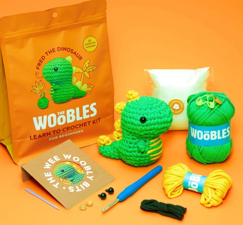 Crochet kit components arranged on an orange background, including an orange branded pouch, a green amigurumi dinosaur, a green yarn ball, yellow yarn, a crochet hook, thread, beads, and a small card. Visible text includes 'THE WOBLES' and 'LEARN TO CROCHET KIT'.