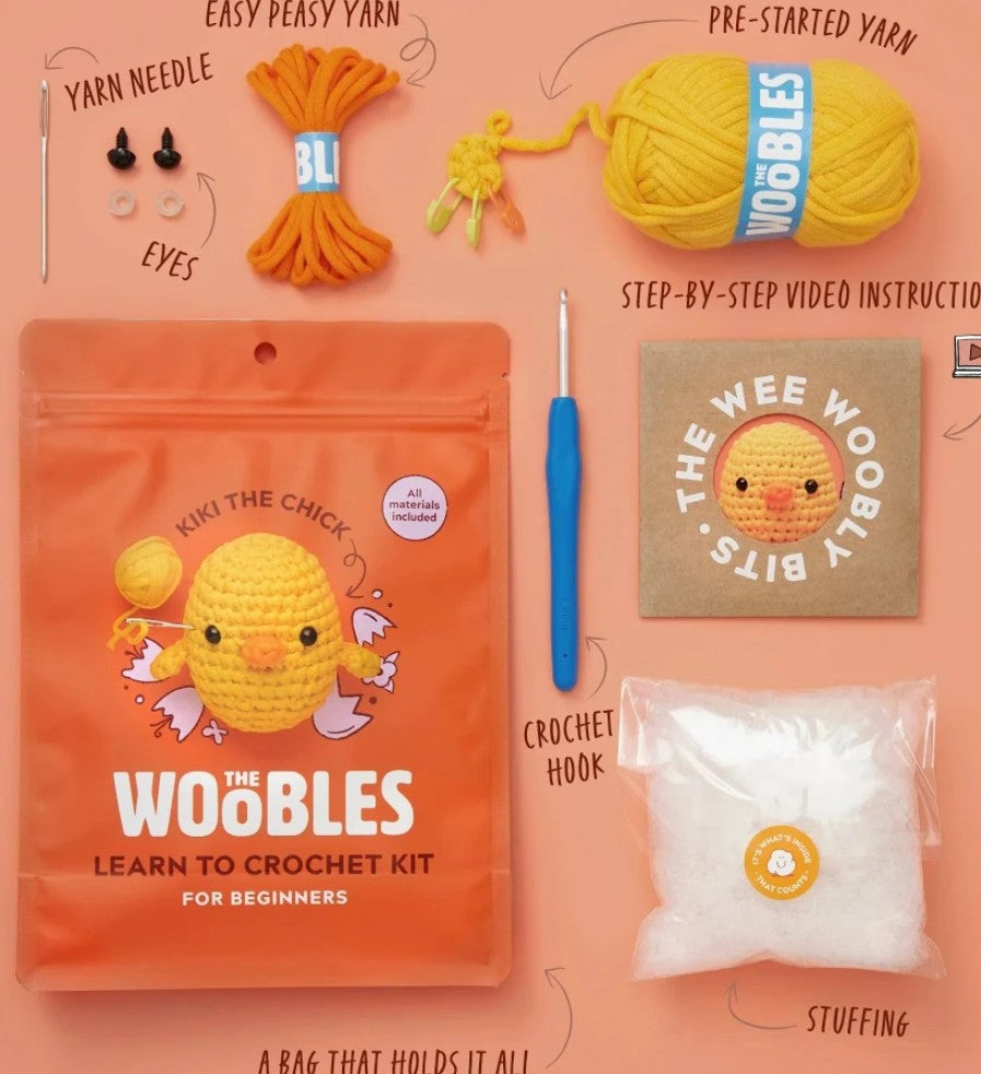 Theee Woobly Bits 'The Woobles' Learn to Crochet Kit for Beginners, featuring an orange pouch with a yellow chick illustration, a yellow ball of yarn, orange yarn, a blue crochet hook, white stuffing, and a cardboard instruction booklet on a peach background with handwritten labels.