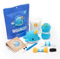 The Woebles Amigurumi Narwhal Crochet Kit displayed on a clean white background, including the blue pouch packaging, a crocheted narwhal, yarn, thread, hook, and accessories.