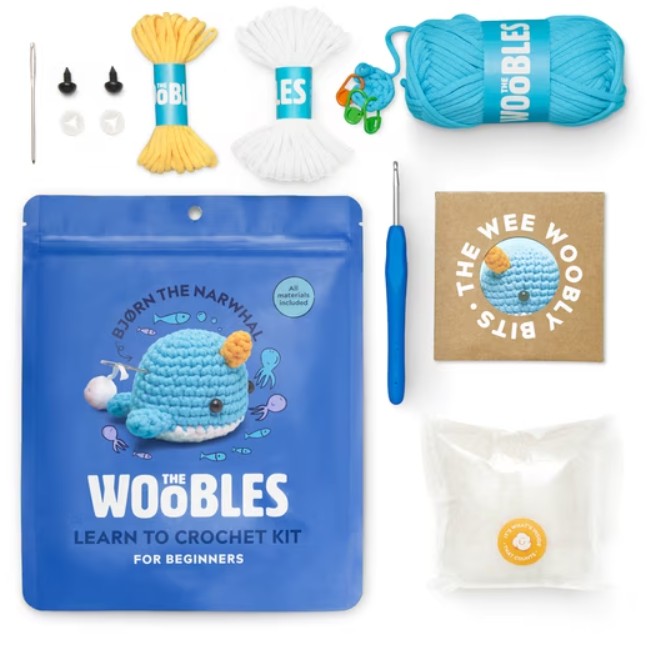 The Woobles Learn to Crochet Kit for Beginners, featuring a blue pouch with a crocheted whale illustration, a blue crochet hook, yellow and white yarn, small black and white beads, a brown card reading 'The WEE Woobly Bits', and a small clear packet, all arranged on a white background.