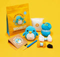 Crochet kit for 'Pierre the Penguin' by Woobles, featuring a yellow pouch, a blue and white crocheted penguin, blue and white yarn, a crochet hook, and other accessories on a bright yellow background.